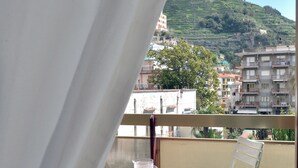 Ein Balkon mit Tisch und Stühlen, Blick auf einen Hügel mit Gebäuden, und ein Vorhang, der das Fenster teilweise verdeckt.