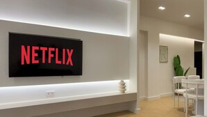 A modern living room with a mounted TV displaying "NETFLIX" in red letters, a white console table, and a seating area with a white chair and a small table. - Elegant Living in Glyfada Center (Glifada)