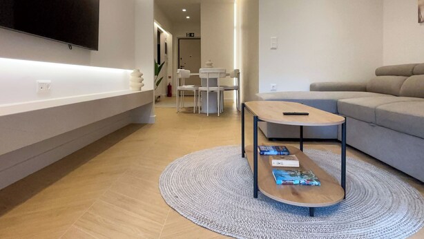 A modern living room with a flat-screen TV mounted on the wall, a grey sofa, a coffee table with books, and a round rug. - Elegant Living in Glyfada Center (Glifada)