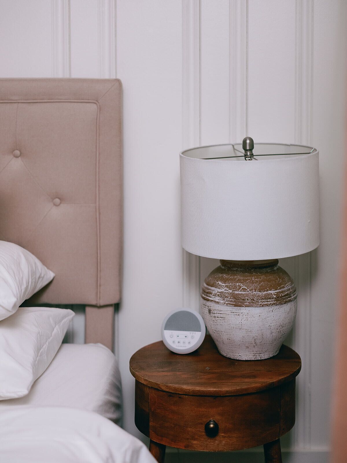A bed with white bedding, a beige headboard, and a bedside table with a lamp and a digital device.
