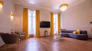 Una sala de estar con piso de madera, un sofá, una silla, un televisor y cortinas amarillas.