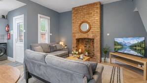 A living room with a grey sofa, a brick fireplace, a television, and a window with blinds. - 4BR Home Perfect for Professionals (Beeston)