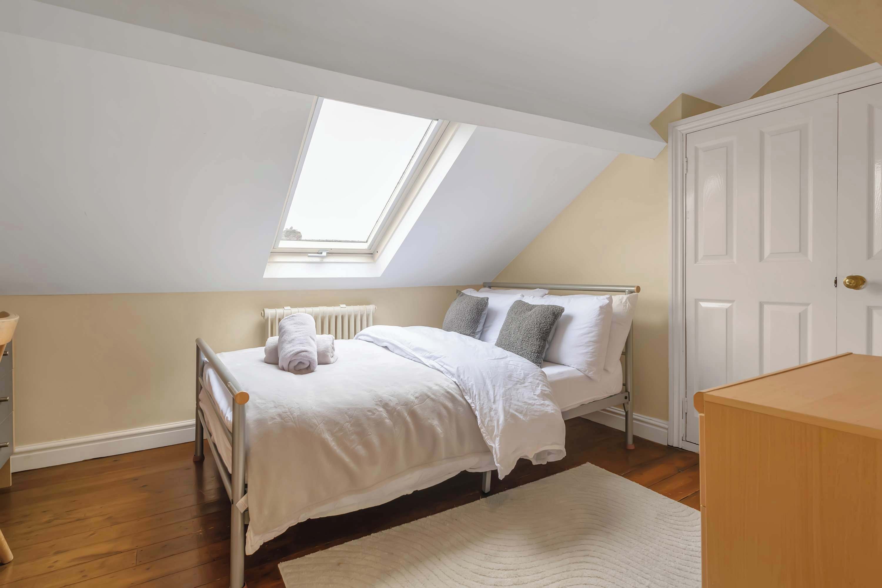 Un dormitorio con tragaluz, cama con ropa de cama blanca, mesita de noche y cómoda de madera.