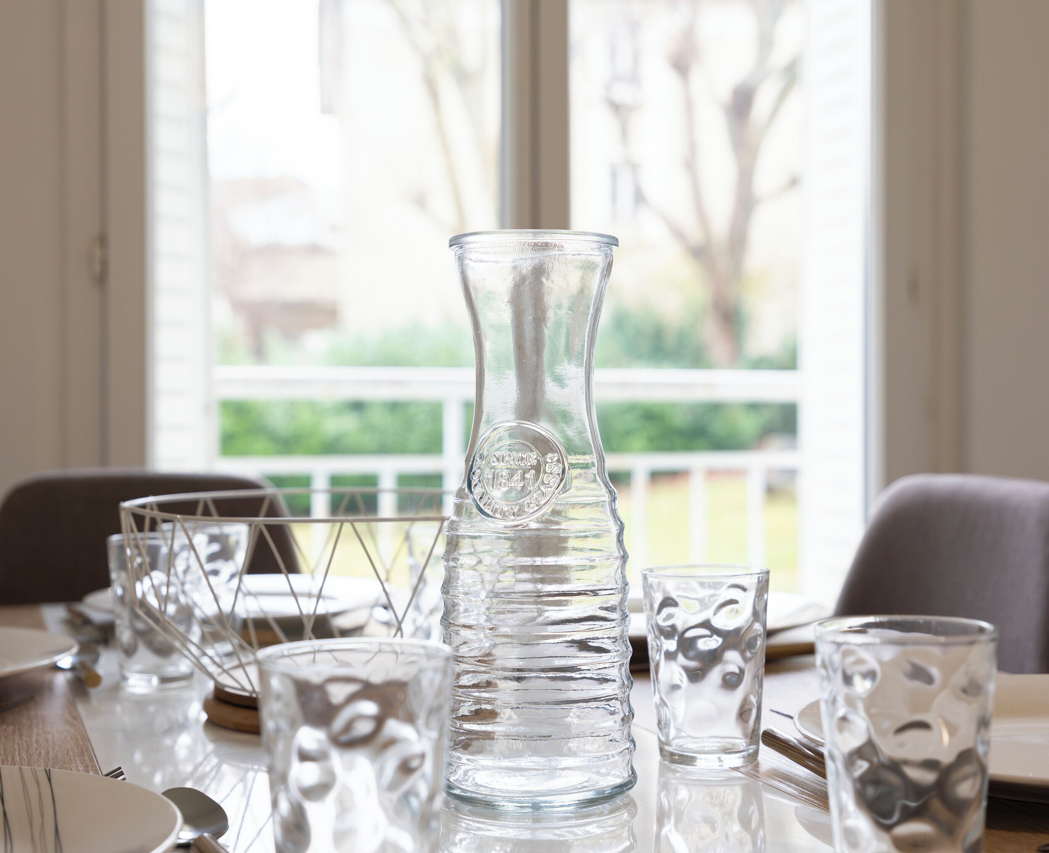 Une table ornée de beaux verres transparents, dont un grand vase et plusieurs verres, est placée devant une fenêtre donnant sur un paysage verdoyant.