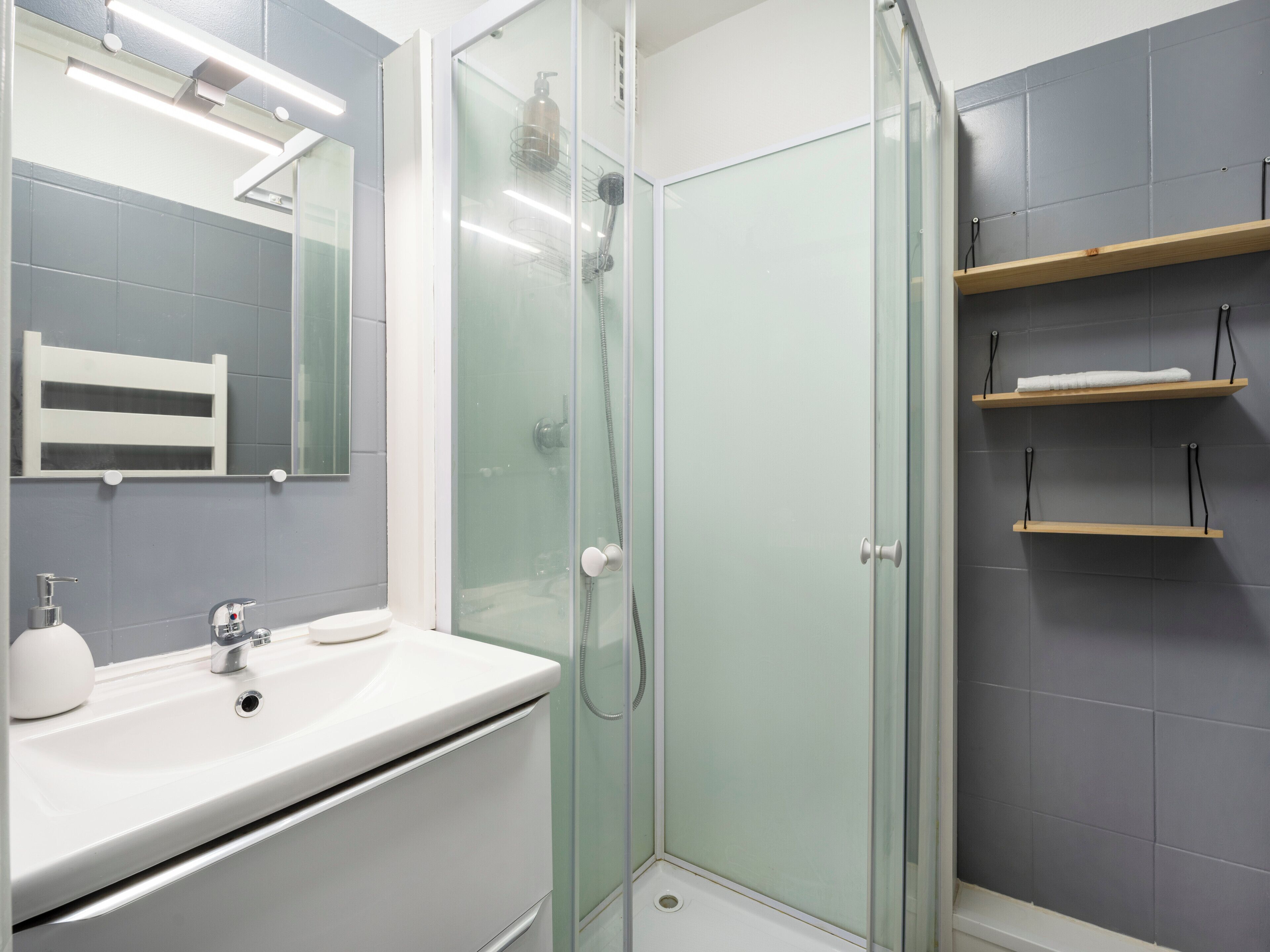 Une salle de bain moderne avec une douche à parois de verre, un lavabo blanc et un miroir au-dessus. Deux étagères en bois sont fixées au mur.
