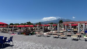 A beachside area with rows of sun loungers and red umbrellas, a gravel ground, and a mountain in the background. - Angelina's House Apartment with Balcony and Air Conditioning (Sant'Anna)