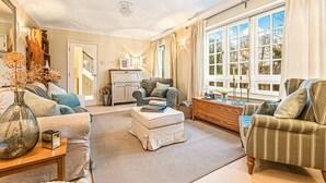 A spacious living room with a sofa, armchair, and ottoman, a wooden coffee table, and a large window with curtains. - Hillside House in Henley (Oxfordshire)