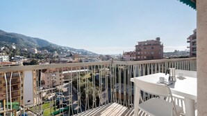 Ein Balkon mit weißem Tisch und Stühlen, mit Blick auf eine Stadtlandschaft mit Gebäuden und einem Berg.