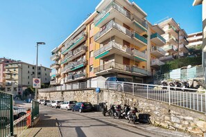 Um edifício residencial com vários andares, varandas, área de estacionamento com carros e motocicletas, e um muro de pedra ao longo da rua.