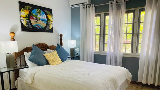Een net opgemaakt bed met witte lakens en blauw-gele kussens, een houten hoofdbord, een nachtkastje met een lamp, en een schilderij van een strandlandschap aan de muur.
