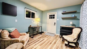 Una habitación con piso de madera, un sofá, una silla, un escritorio con una lámpara, una puerta y una estantería en la pared.