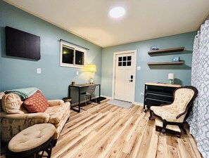 A room with a wooden floor, a sofa, a chair, a desk with a lamp, a door, and a wall-mounted shelf. - Downtown Boone / 30+ Nights / 4BR Home (Boone)