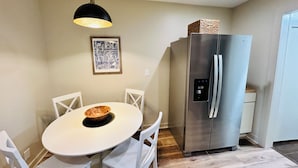 Cocina compacta con una mesa blanca, cuatro sillas, un refrigerador de acero inoxidable y una lámpara colgante.