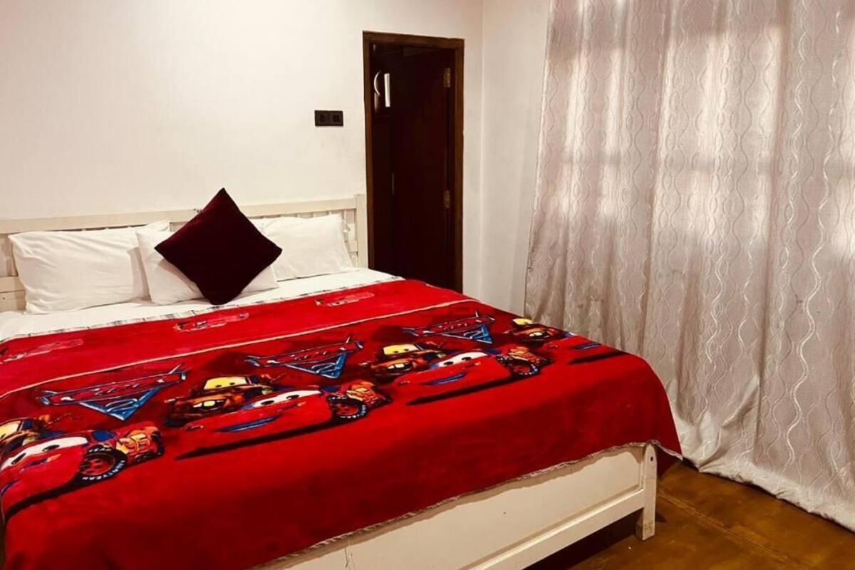Une chambre à coucher avec un lit, une tête de lit blanche, une couverture rouge à motifs de voitures, des rideaux blancs et une porte en bois.