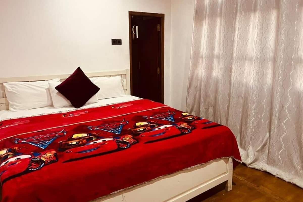 A bedroom with a bed, white headboard, red blanket with car prints, white curtains, and a wooden door.