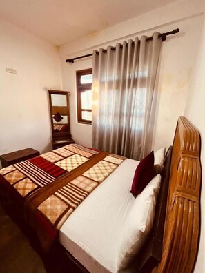 A bedroom with a bed, a wooden headboard, a nightstand, a mirror, and a window with curtains. - The Budget White House Nuwaraelliya (Nuwara Eliya)