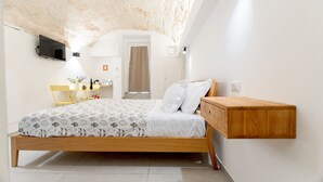 Un dormitorio con una cama de madera, un televisor montado, un escritorio con una silla y una mesita con un florero.