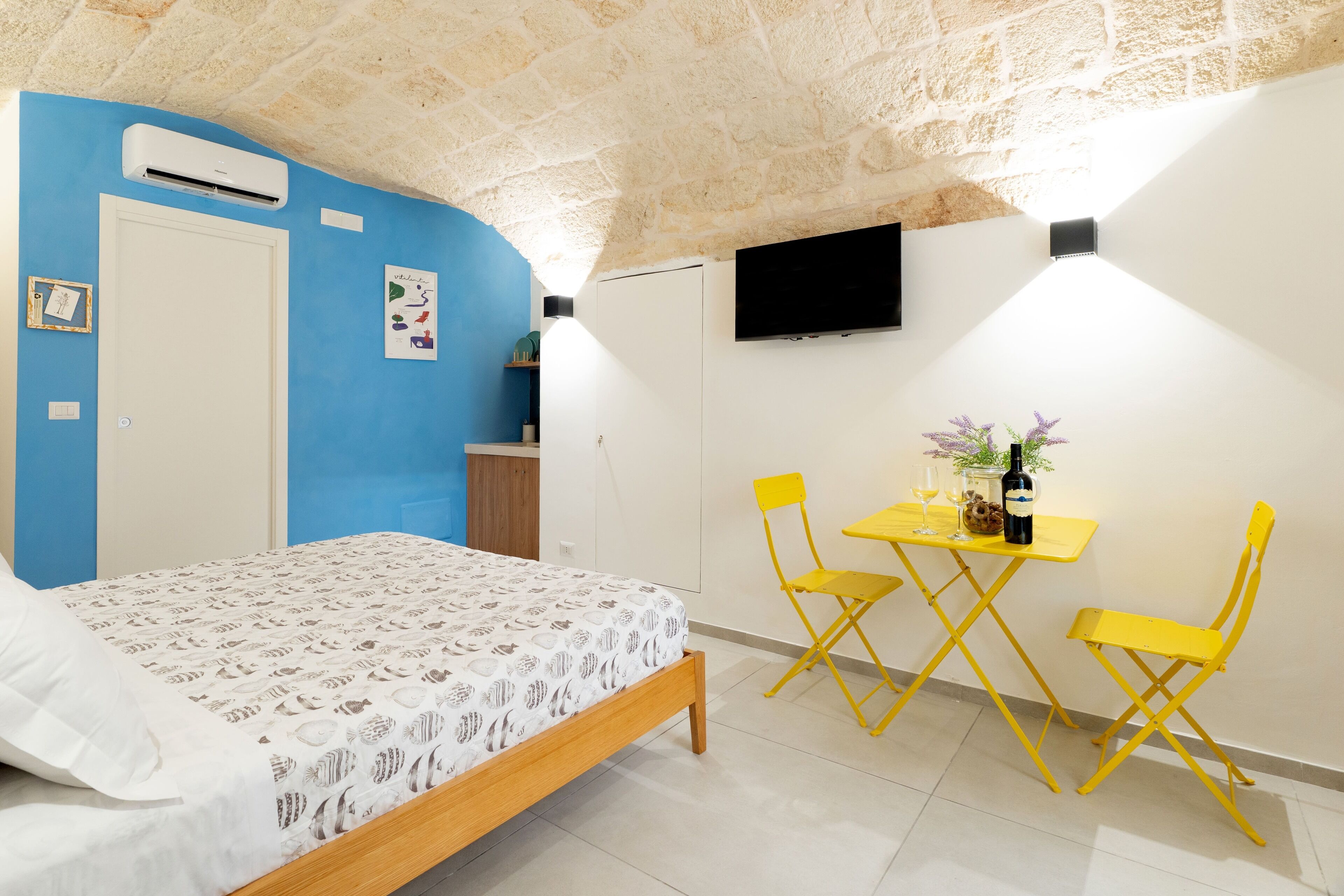 Un dormitori amb un llit, una paret blava, una porta blanca, una televisió de pantalla plana i una petita zona de menjador amb cadires grogues i una taula.