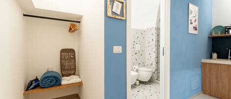 A bathroom with a blue wall, a white toilet, a shower area, and a shelf with towels and a folded blanket.