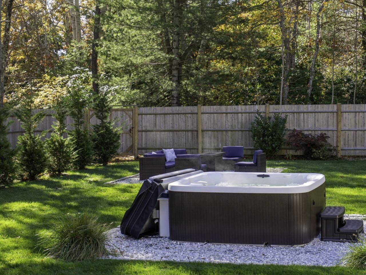 Un jacuzzi rodeado de mobiliario exterior en un patio trasero con valla de madera y árboles.