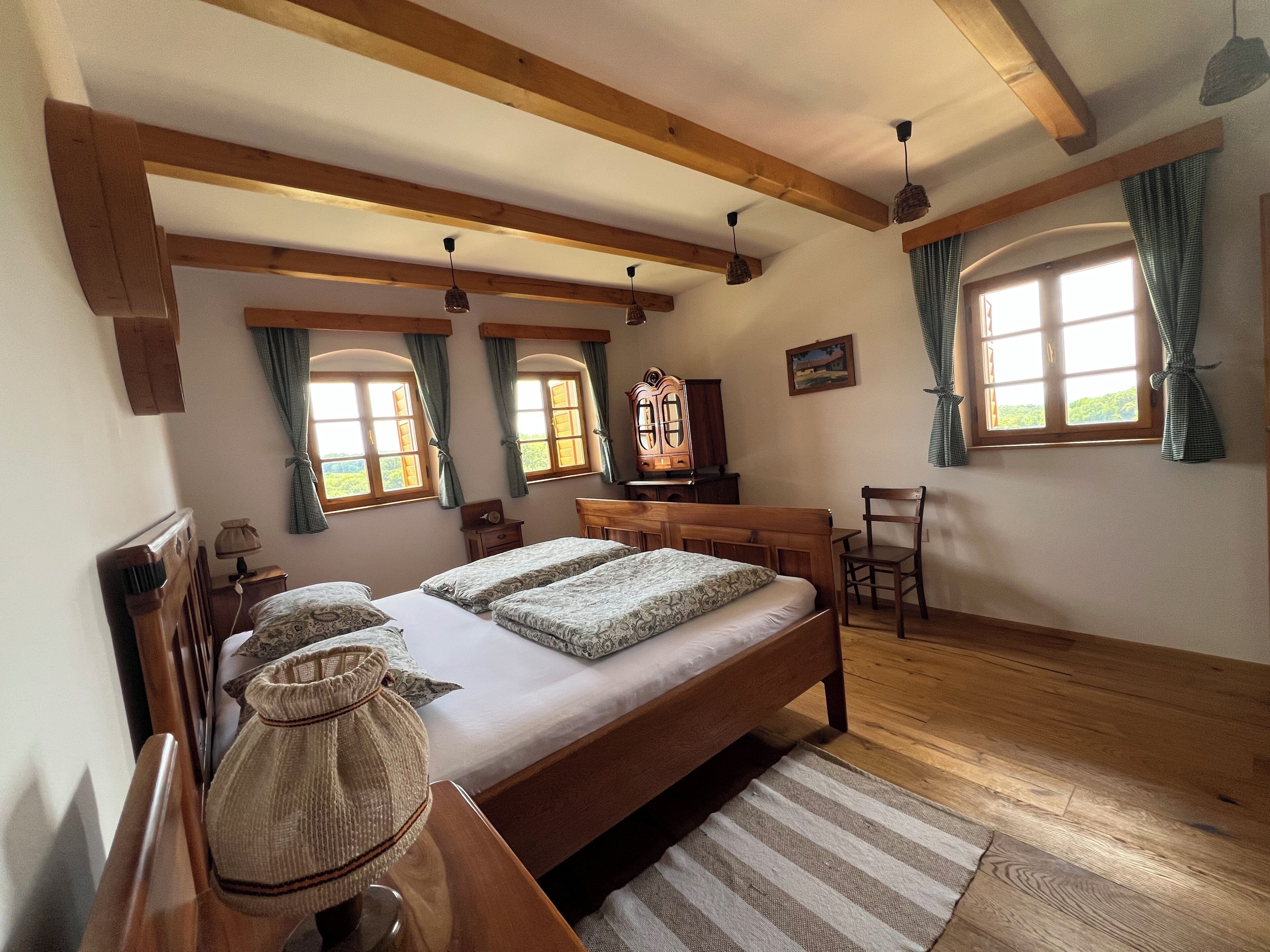 Ein Zimmer mit einem großen Bett, Holzbalken, einem Schreibtisch und zwei mit Vorhängen versehenen Fenstern.