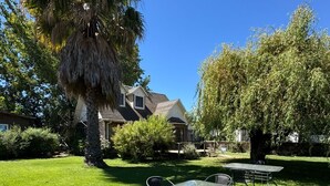 Un césped cuidado con una mesa y sillas, un árbol grande y una casa al fondo.