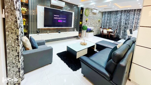 A modern living room with a flat-screen TV, grey sofas, a coffee table, and a dining area with a table and chairs. - Alubarika Service Apartments (Lagos)