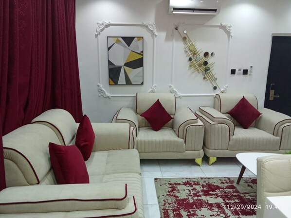 A living room with two beige sofas, red cushions, a geometric abstract painting, a decorative wall piece, and a red and white patterned rug.