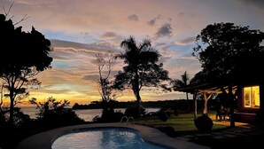 Une piscine au crépuscule, offrant une vue sur le coucher de soleil se reflétant sur une étendue d’eau, entourée d’arbres et d’un petit bâtiment dont les lumières sont allumées.