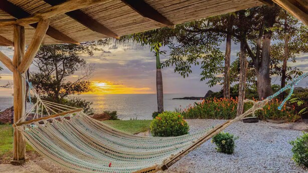 Un hamac sous une pergola en bois, avec vue sur une plage au coucher du soleil.