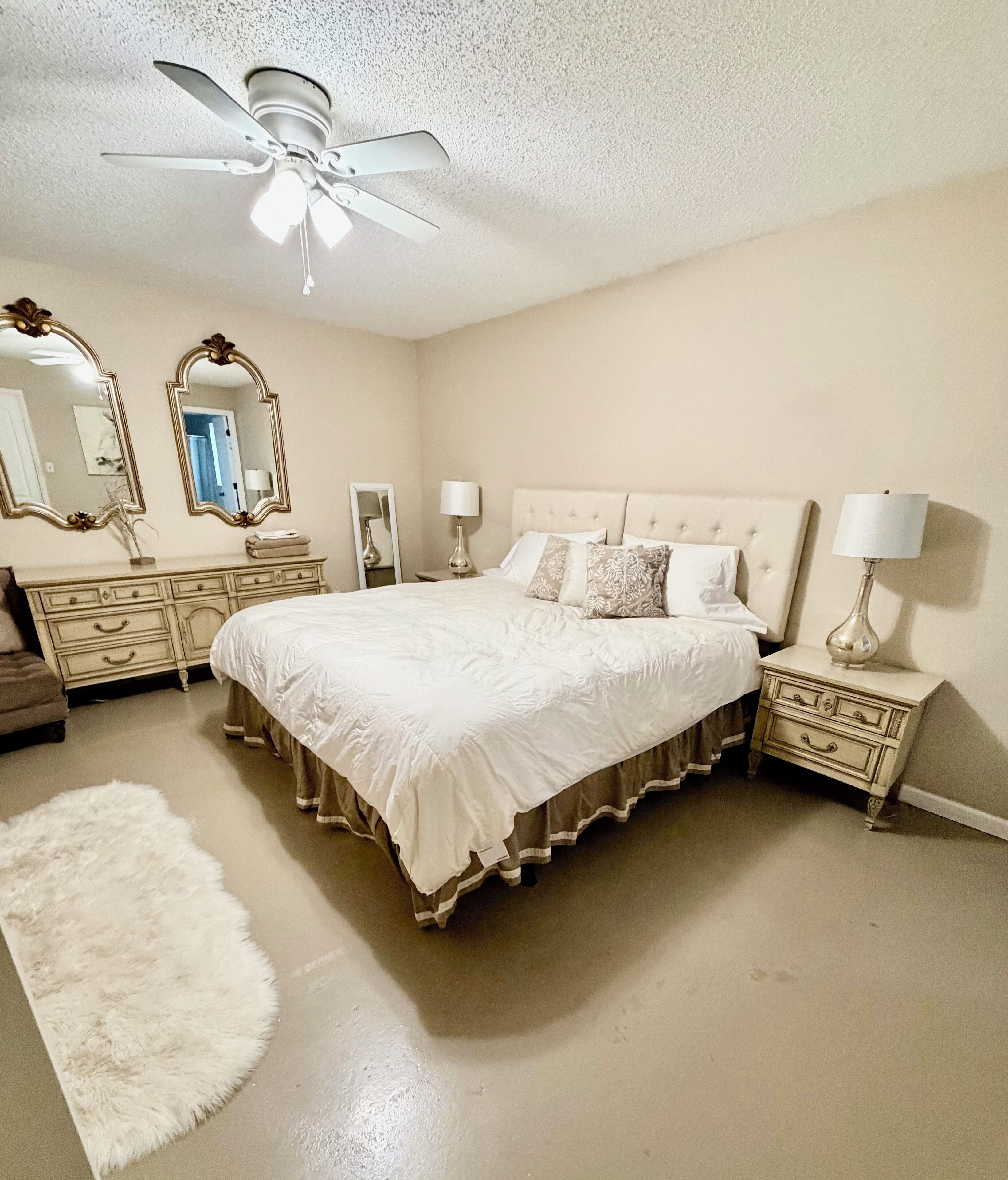 A bedroom with a large bed, two nightstands, a dresser, a ceiling fan, and two wall mirrors.
