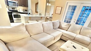 A modern living room with a sectional sofa, a small ottoman, and a kitchen area with appliances.