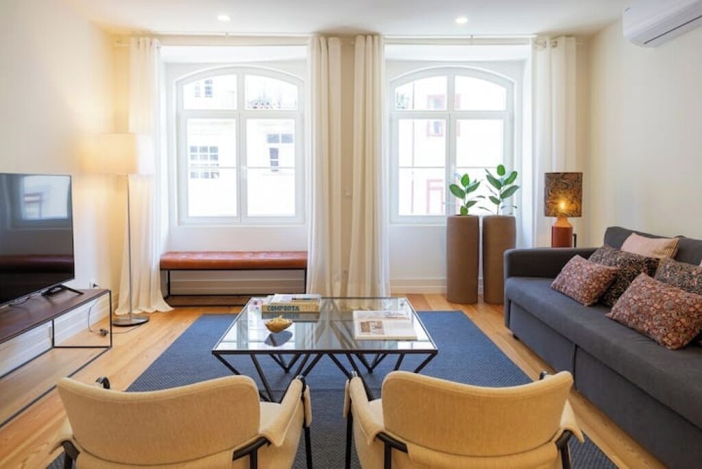 Ein modernes Wohnzimmer mit einem Glastisch, zwei beigefarbenen Sesseln, einem dunklen Sofa mit gemusterten Kissen, einem Fernseher auf einem Schrank und großen Fenstern mit Vorhängen.