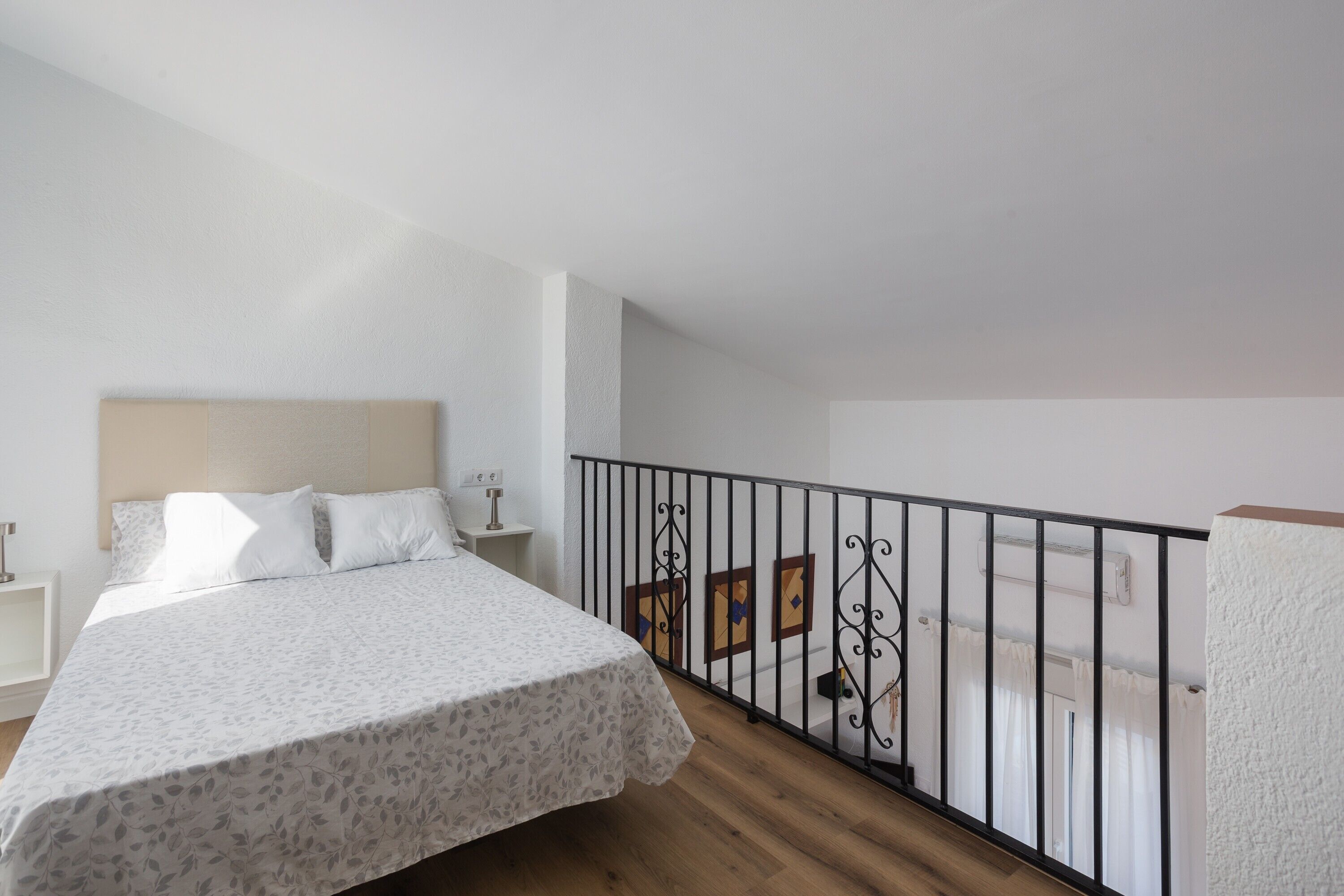 A bedroom with a bed, a nightstand, a railing, and a view of the outside.