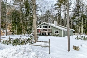 Ein zweistöckiges Haus mit Balkon, umgeben von verschneiter Landschaft und hohen Bäumen.