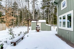 Schnee bedecktes Gelände mit einer Holzbank, einem kleinen Schuppen und einer Pergola.