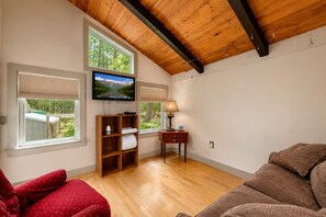 Ein Zimmer mit Holzboden, einem an der Wand befestigten Fernseher, einem Holzkasten mit Blick aus dem Fenster, einem Schreibtisch mit Lampe und einem roten Sofa.