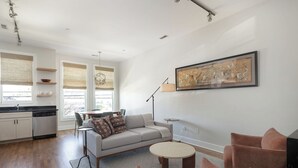 Una sala moderna con un sofá, dos sillones, una mesita de centro y un comedor con mesa y sillas. Cuenta con una zona de cocina con armarios y lavavajillas. Un cuadro grande enmarcado adorna la pared, y la habitación tiene ventanas amplias con persianas.
