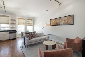 A modern living room with a sofa, two armchairs, a coffee table, and a dining area with a table and chairs. There is a kitchen area with cabinets and a dishwasher. A large framed artwork is on the wall, and the room has large windows with blinds. - 12 South Townhome across from Sevier Park (Nashville)