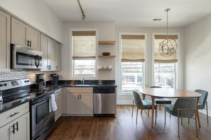 A modern kitchen with a stainless steel oven, microwave, and dishwasher, wooden cabinets, and a dining area with a round table and chairs. - 12 South Townhome across from Sevier Park (Nashville)
