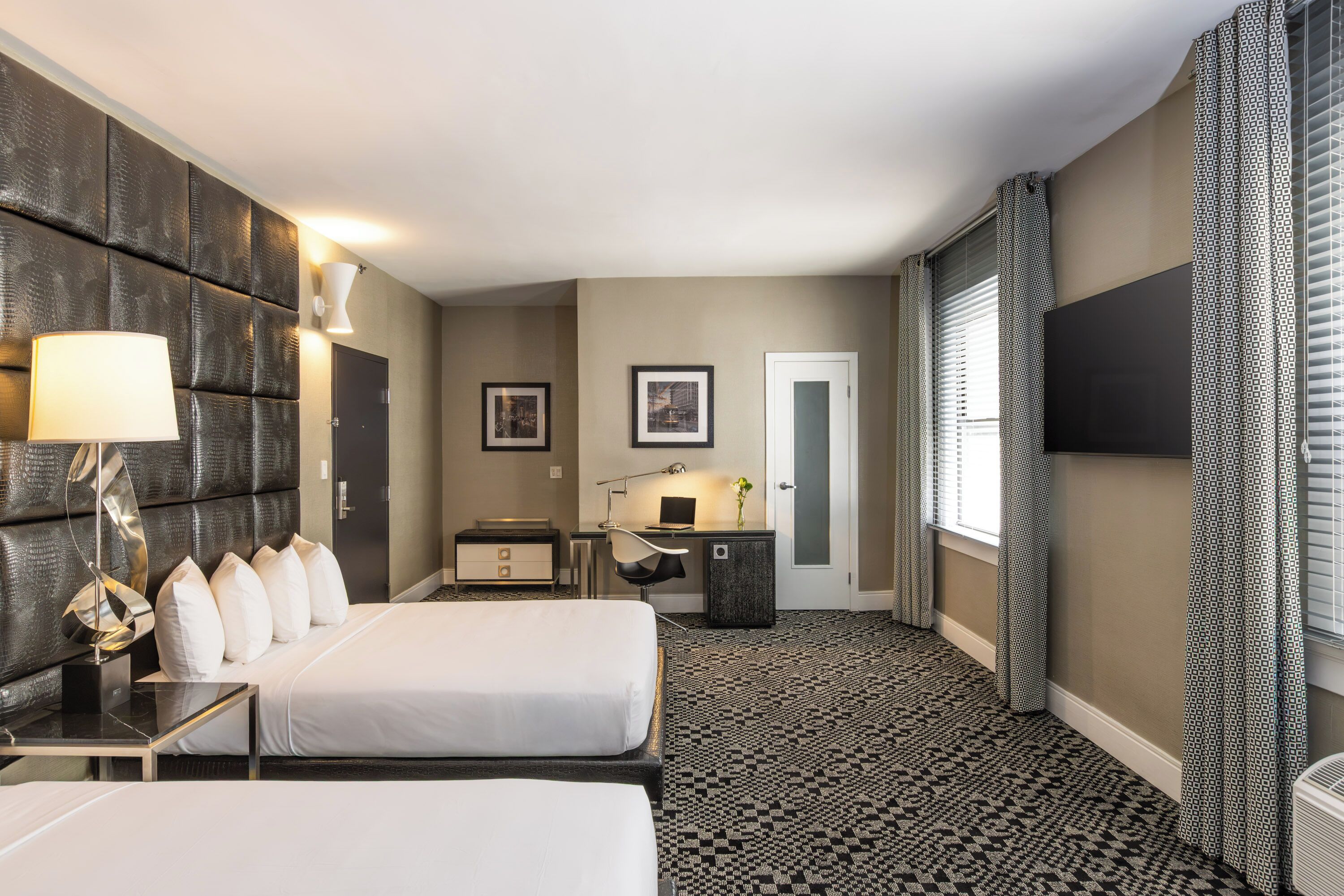 Ein Hotelzimmer mit zwei Betten, einem Schreibtisch, einem Stuhl, einem kleinen Tisch, einer Lampe, einem Bilderrahmen, einem Spiegel, einem Fenster mit Vorhängen und einem an der Wand montierten Fernseher.