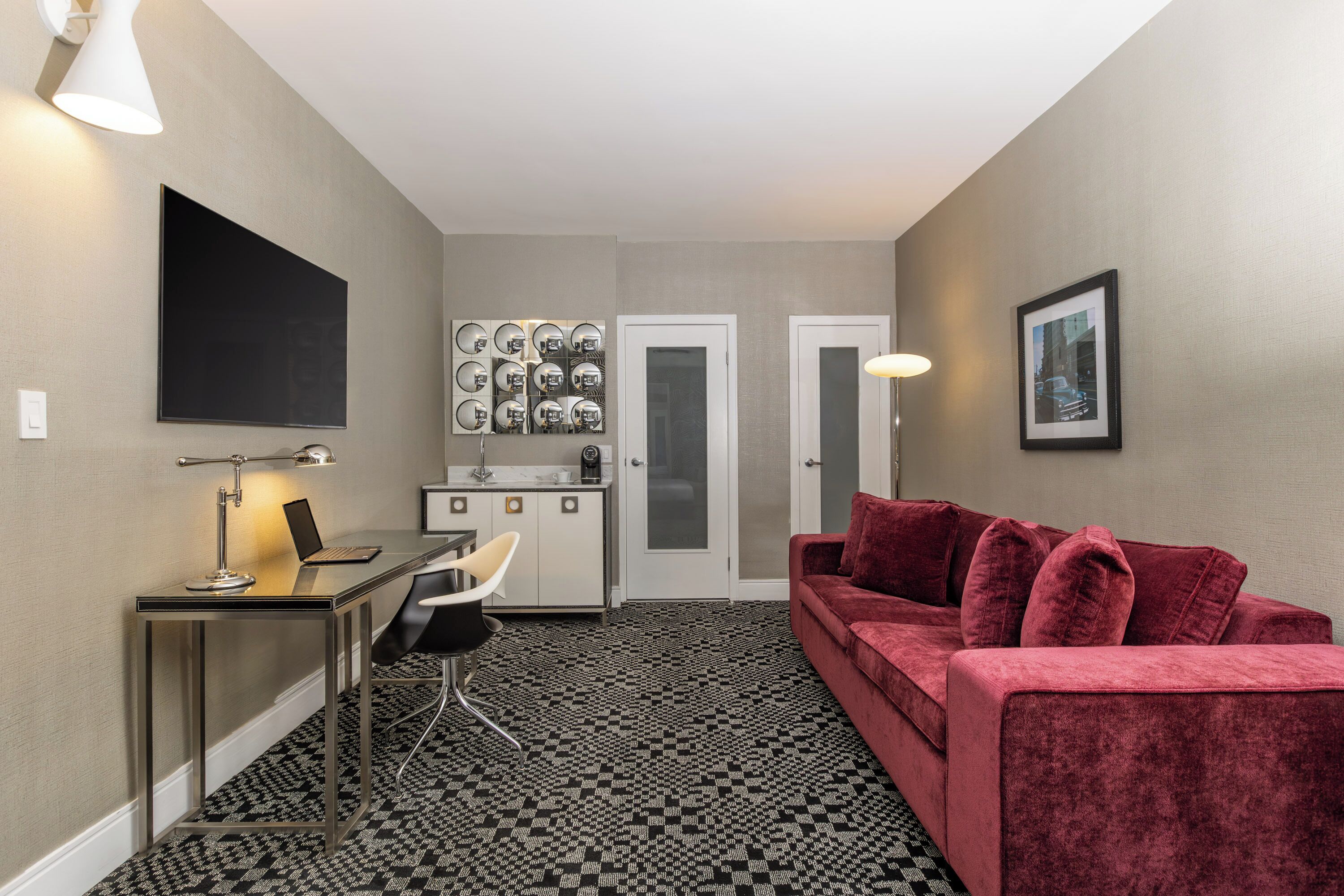 A compact living space with a kitchenette, a seating area with a red sofa, a flat-screen TV mounted on the wall, and a patterned floor.
