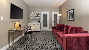 A compact living space with a kitchenette, a seating area with a red sofa, a flat-screen TV mounted on the wall, and a patterned floor.