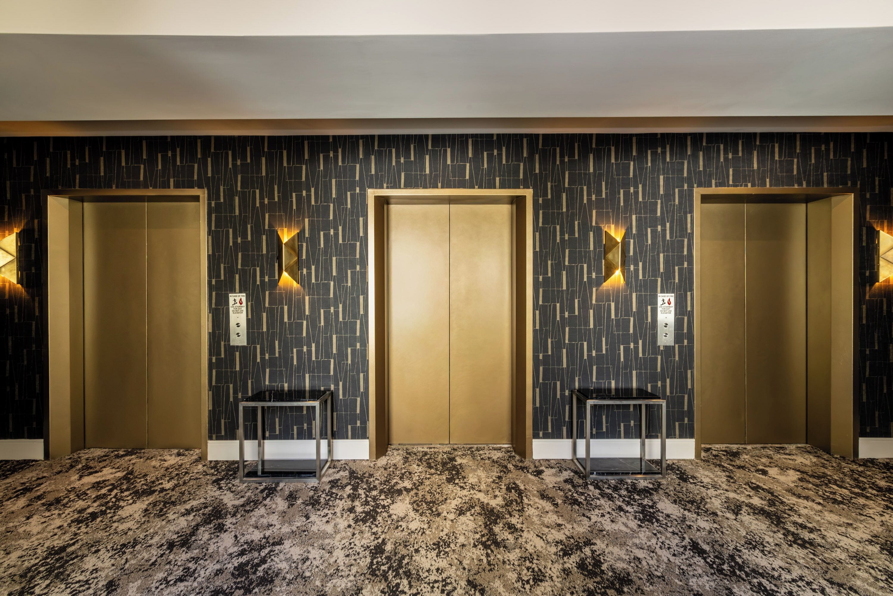 A hotel lobby with three elevators, a patterned wall, and a carpeted floor.