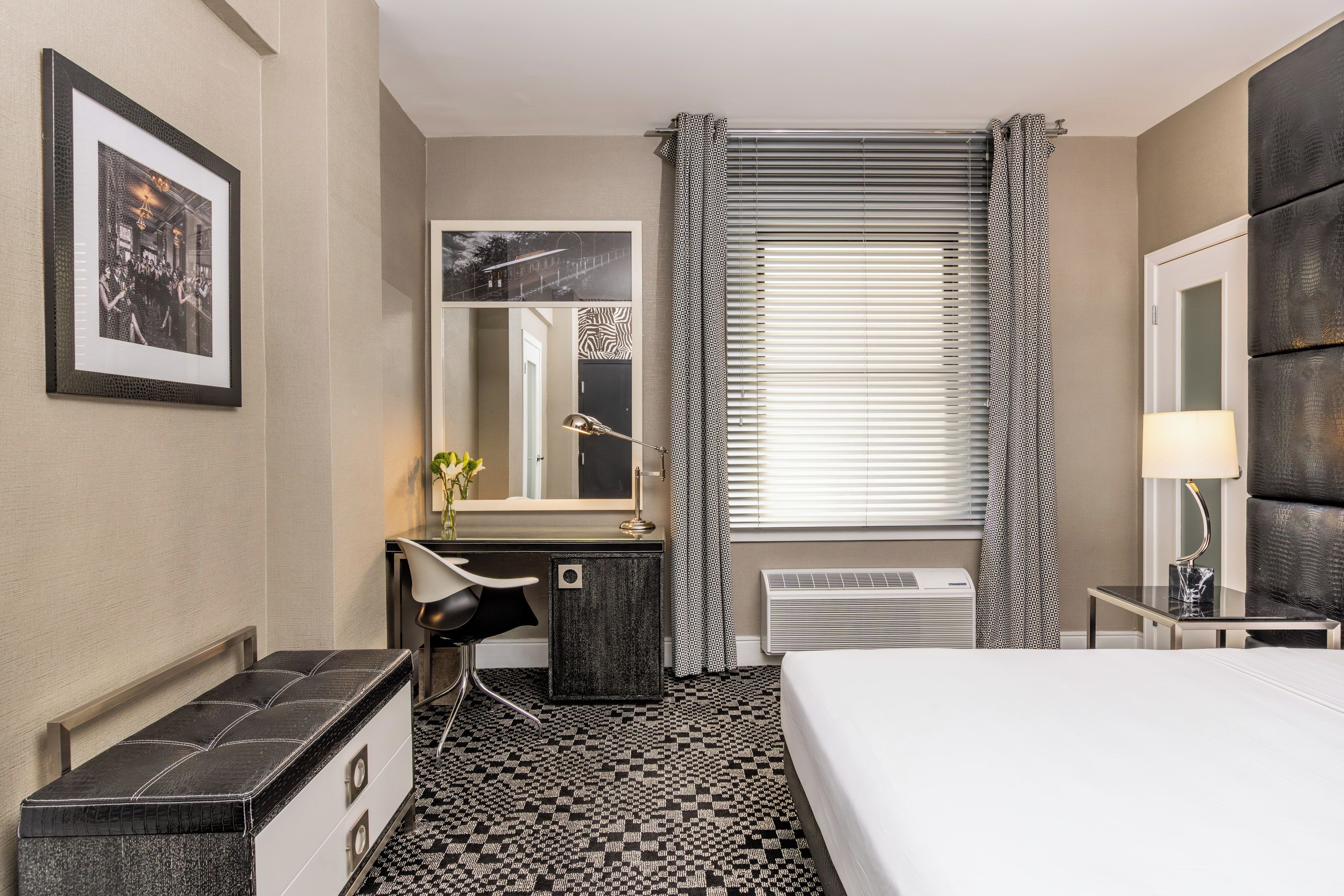 A hotel room with a bed, a desk with a chair, a mirror, a window with blinds, and a radiator.