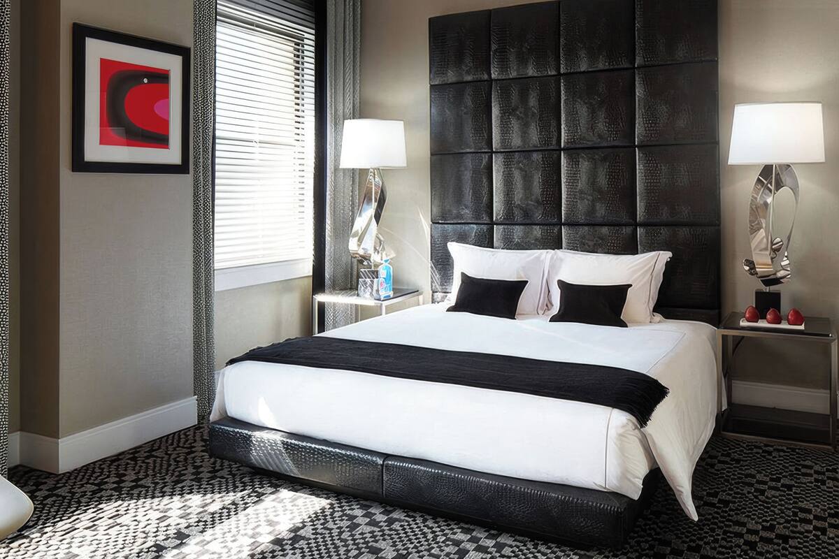 A hotel room with a large bed, two bedside tables with lamps, a headboard, a framed artwork, and a patterned carpet.