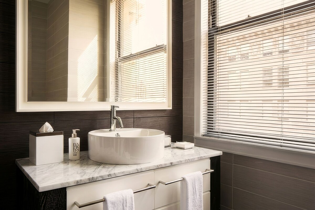 A bathroom with a large mirror, a round sink, and a window with blinds.