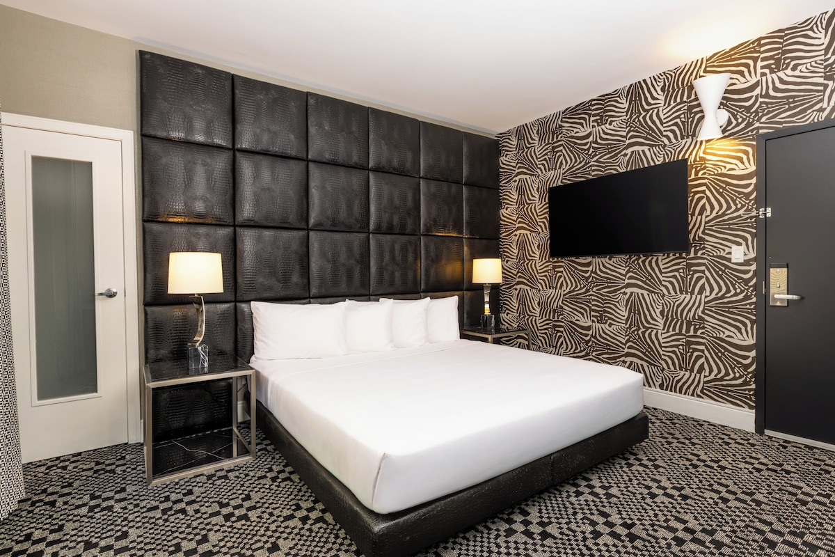 A hotel room with a large bed, two bedside tables with lamps, a flat-screen TV mounted on the wall, and a zebra-patterned wallpaper.