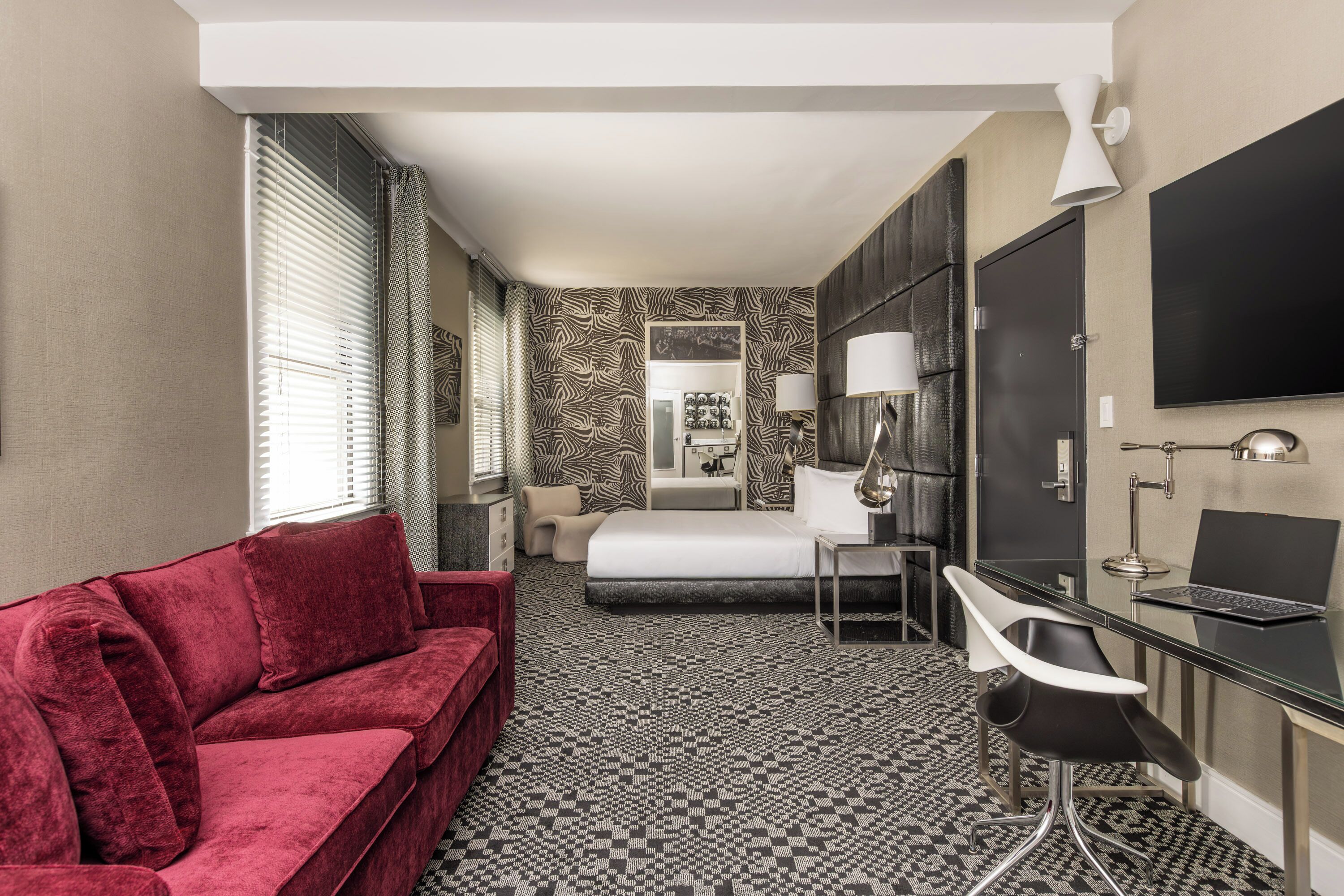 A hotel room with a red sofa, a bed, a desk with a chair, a flat-screen TV, and a patterned carpet.