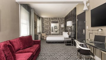 A hotel room with a red sofa, a bed, a desk with a chair, a flat-screen TV, and a patterned carpet.